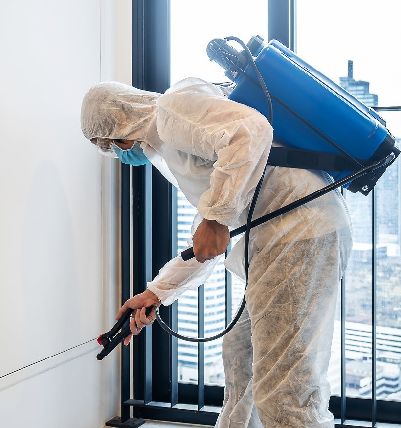 A technician in protective coveralls treating a baseboard beside a window